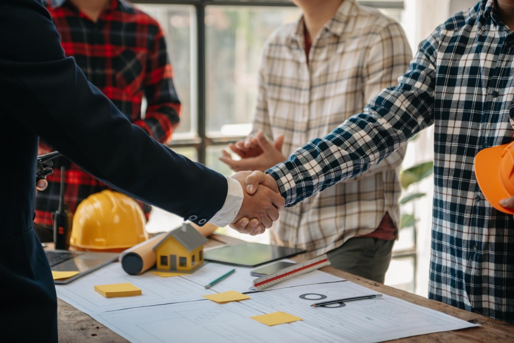 Two people are shaking hands over a table with architectural plans, safety helmets, and a model house, indicating a professional agreement or collaboration.