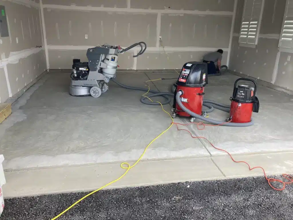 An empty garage with a concrete floor features a floor grinding machine and two vacuum cleaners with hoses and cables sprawling across the surface.