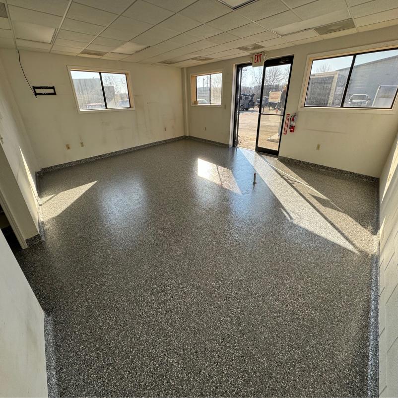 The image shows an empty room with speckled flooring, white walls, and sunlight coming through the windows, creating shadows on the floor.