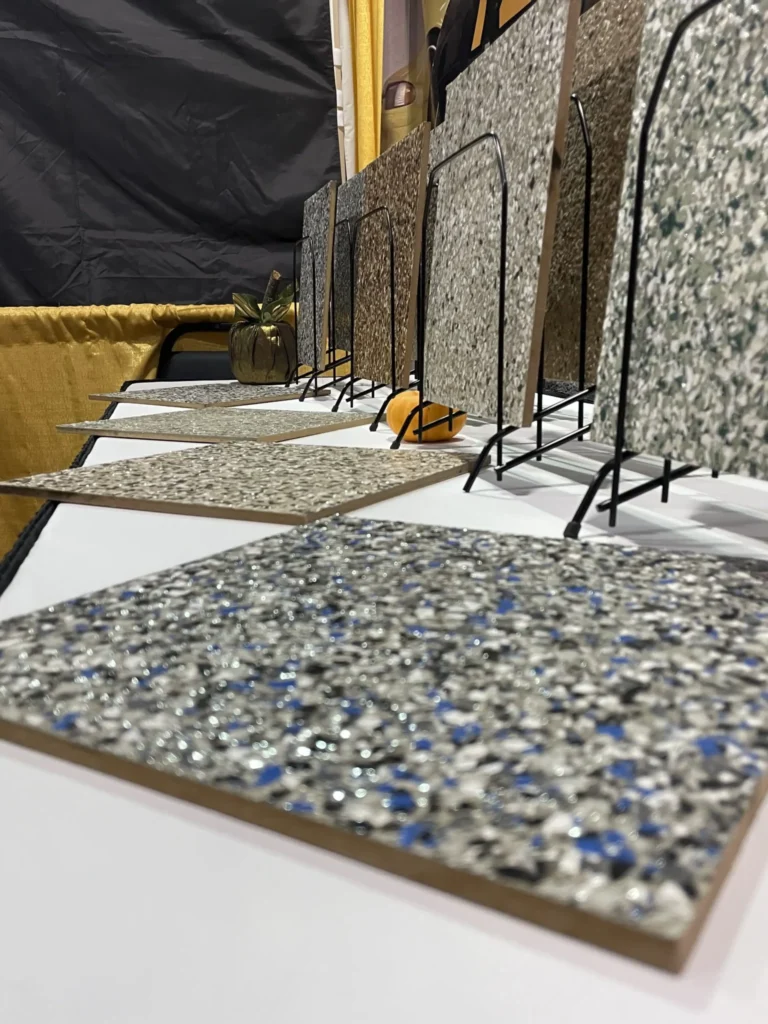 A display of various textured tiles arranged in a vertical rack against a backdrop of black and gold fabric, featuring a small potted plant.