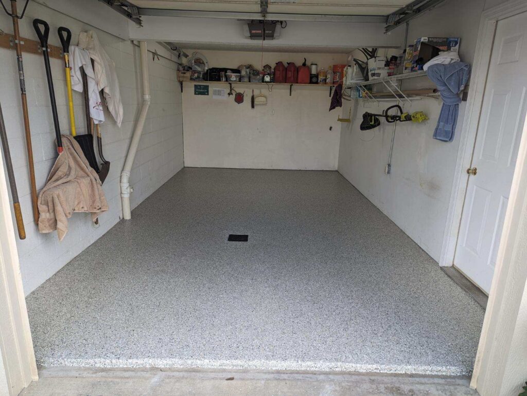 A clean, empty garage with tools and supplies on shelves and walls, featuring a speckled floor and an open garage door.