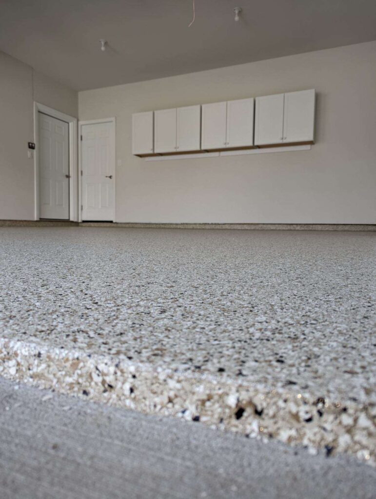 An empty garage with speckled flooring, white cabinets on the wall, and a closed door. The walls are light grey.