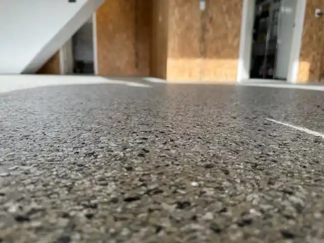 Close-up view of a speckled concrete floor in an indoor space. Wooden-paneled walls and an open doorway are visible in the background.