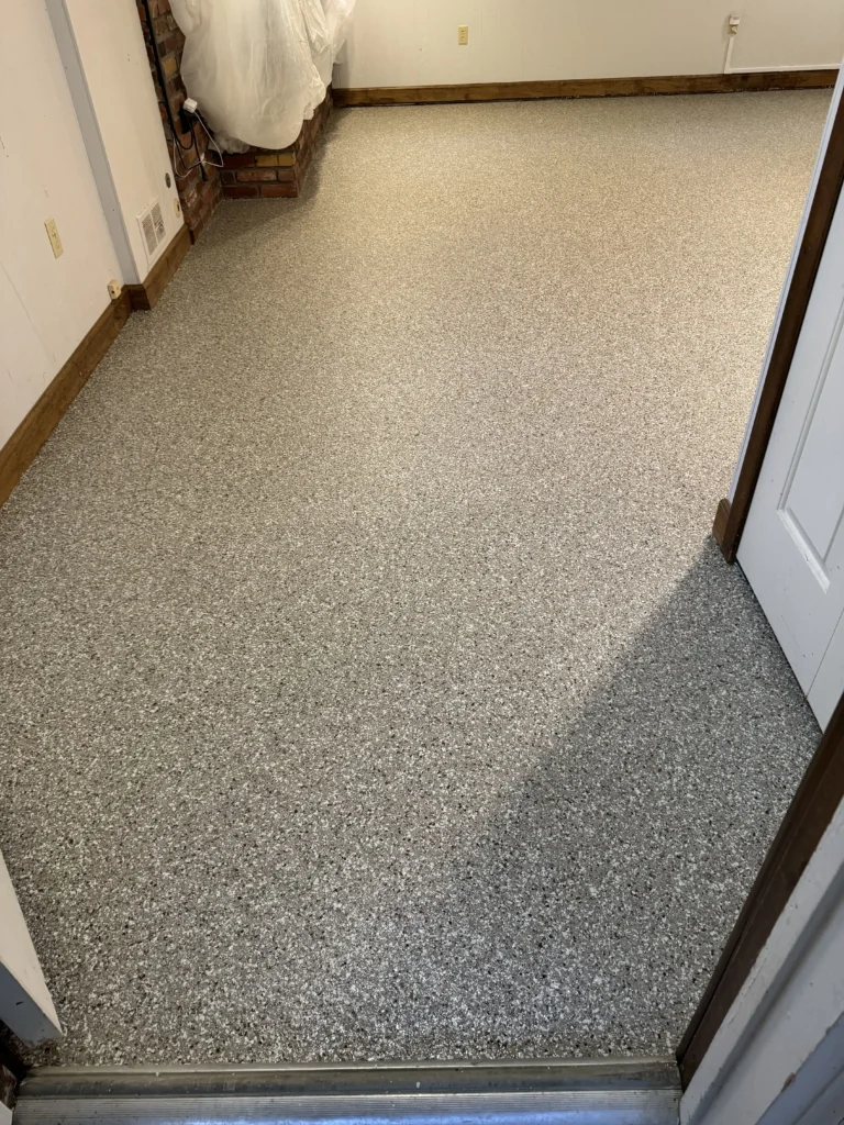 A room with speckled flooring, beige walls, and a wrapped object near a brick corner. Wooden baseboards line the perimeter.