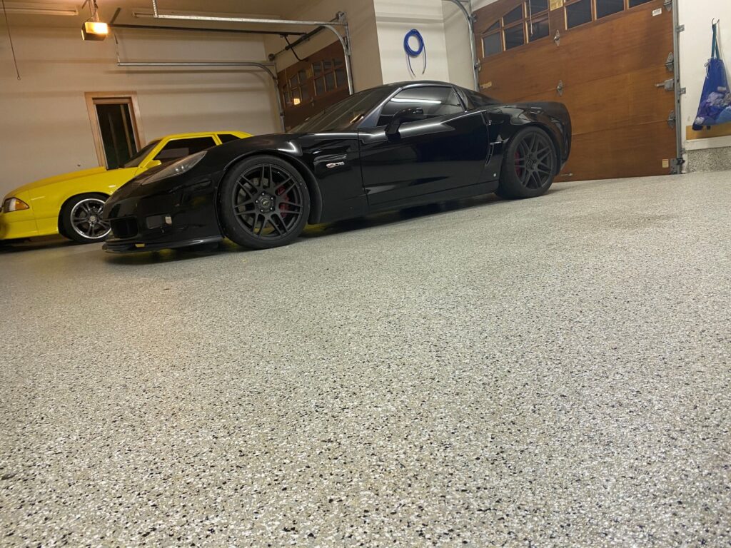 A garage with a black sports car in the foreground and a yellow car in the background, surrounded by clean walls and a concrete floor.