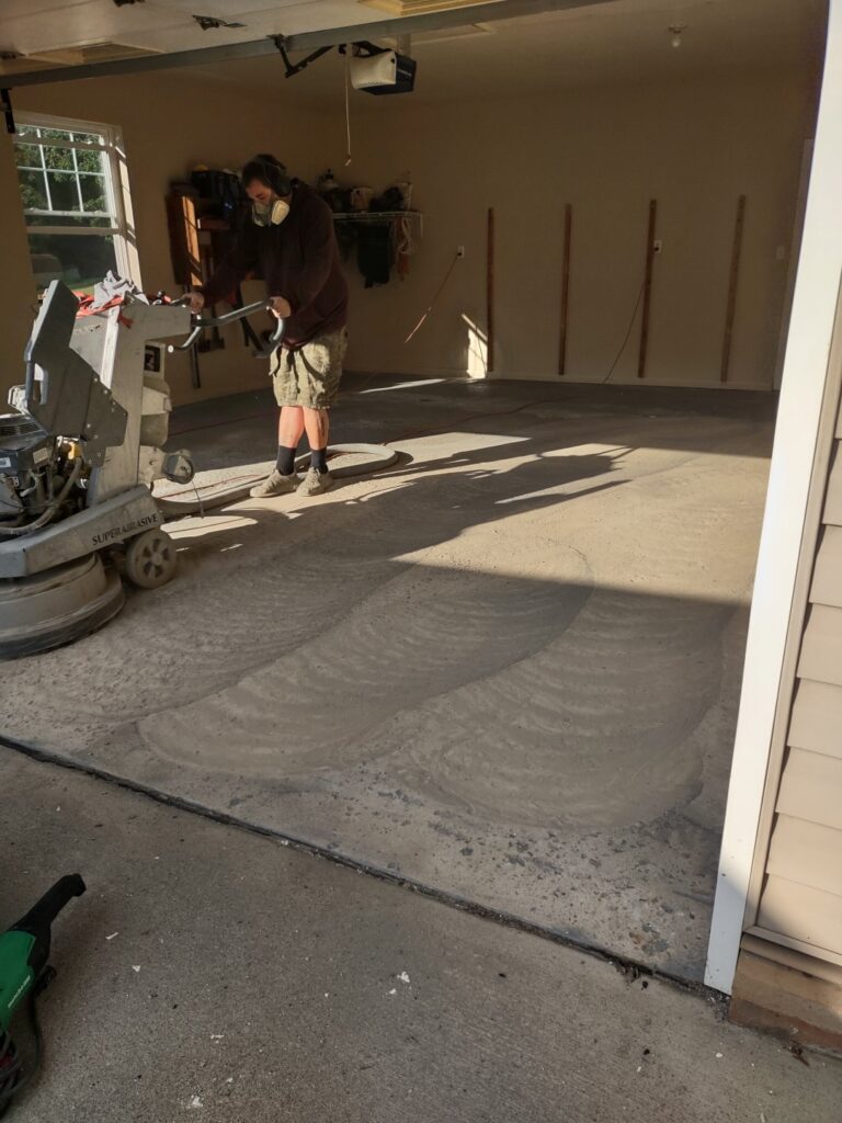 A person operates a floor grinding machine inside a garage, smoothing the concrete floor. Sunlight casts shadows on the unfinished surface.