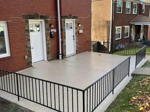 The image shows a concrete patio with a black metal railing in front of a red brick duplex with identical white doors and autumn leaves scattered around.