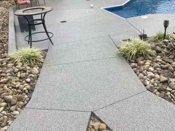 A backyard with a modern gray textured patio leading to a pool. There's outdoor furniture, decorative rock bed, and green plants around the area.