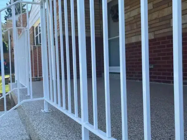 A white metal security gate installed in front of a house's porch with a glass door, red brickwork, and a textured concrete walkway visible.