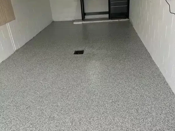 A clean, empty garage with a speckled epoxy floor coating. White painted walls and a central floor drain. Storage cabinets and a workbench at the back.