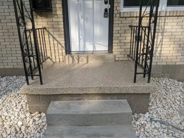 This image shows a residential entrance with a concrete step leading to a textured porch flanked by black wrought-iron railings, with a white rock garden beside it.