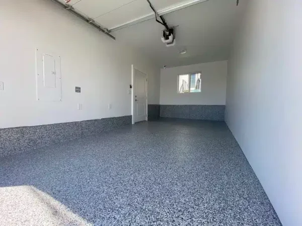 An empty garage with a speckled epoxy floor, light gray walls, and baseboard. A garage door, one window, and entry door are visible.