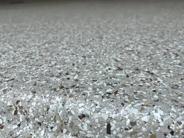 The image shows a close-up of a speckled epoxy floor with scattered flakes, reflecting light, with the blurred background of a vacuum cleaner and wall socket.