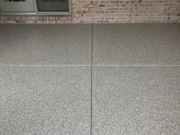 The image shows a textured concrete floor leading up to a building's exterior with a brick wall and glass doors, indicating an entrance or patio.