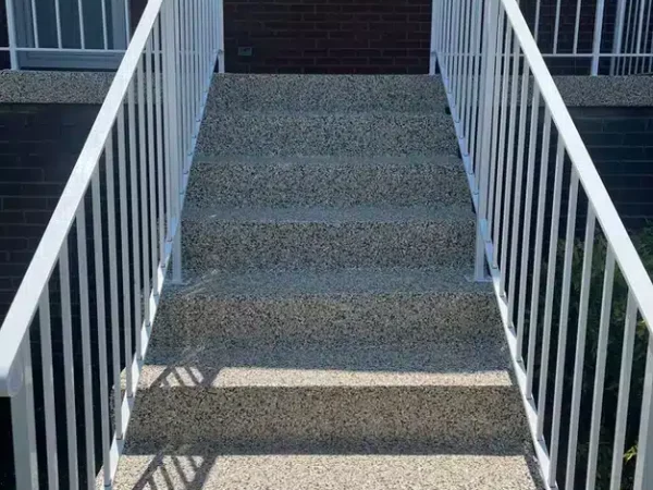 A pebble-textured staircase flanked by white metal railings leads up to a brick building with a door and windows, casting shadow stripes in sunlight.