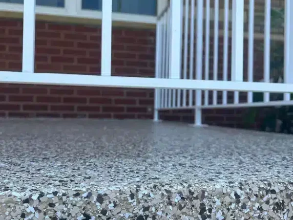 The image displays a close-up of a textured, speckled concrete step with white railing, leading up to a red brick building with closed windows.