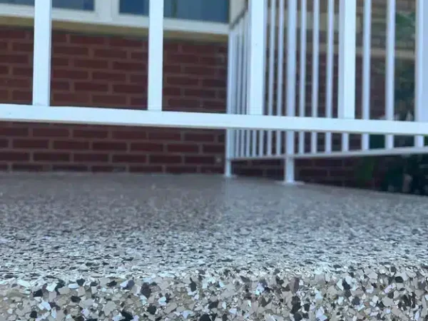 The image displays a close-up of a textured, speckled concrete step with white railing, leading up to a red brick building with closed windows.